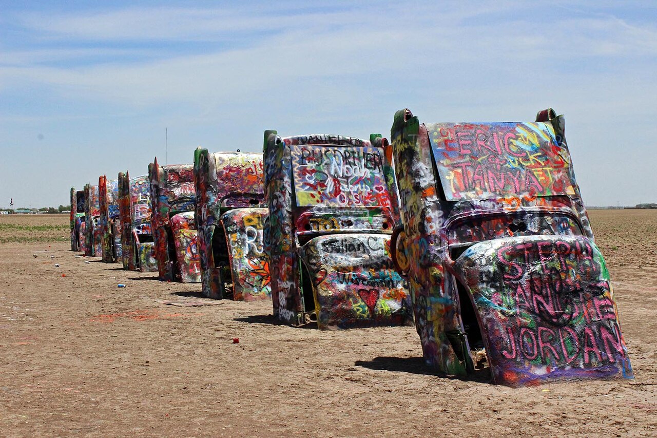 Row of Cadillacs buried nose-first at an angle in a Texas field, their tailfins in the air and their bodies covered in layers of spray-paint graffiti, under a wide sky.
