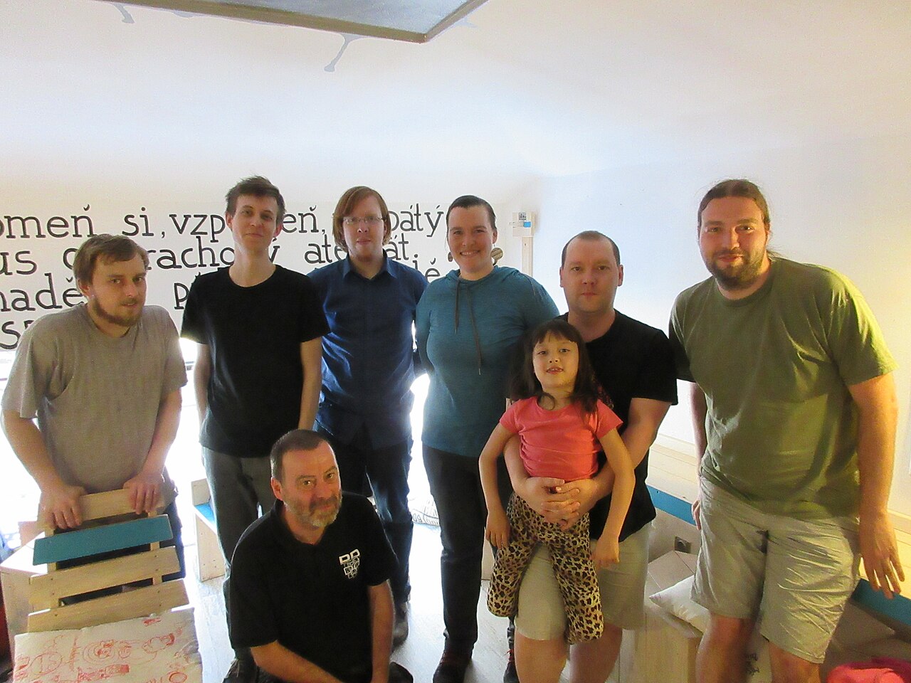 A group of eight people posing together indoors in front of a wall of handwritten Czech text.
