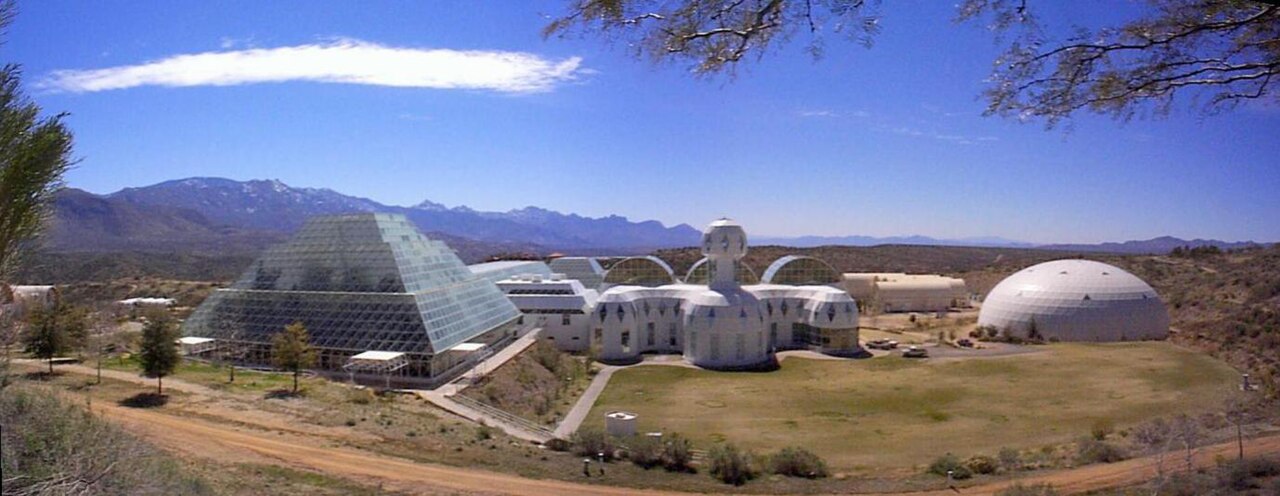 Wide exterior view of the Biosphere 2 complex near Tucson, Arizona: white steel space-frame greenhouses and a stepped glass pyramid rising from the desert.