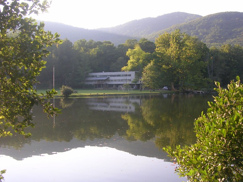 A low modern building reflected in a still lake at dusk, ringed by trees and mountains — the later main building on the Lake Eden campus, now Camp Rockmont.
