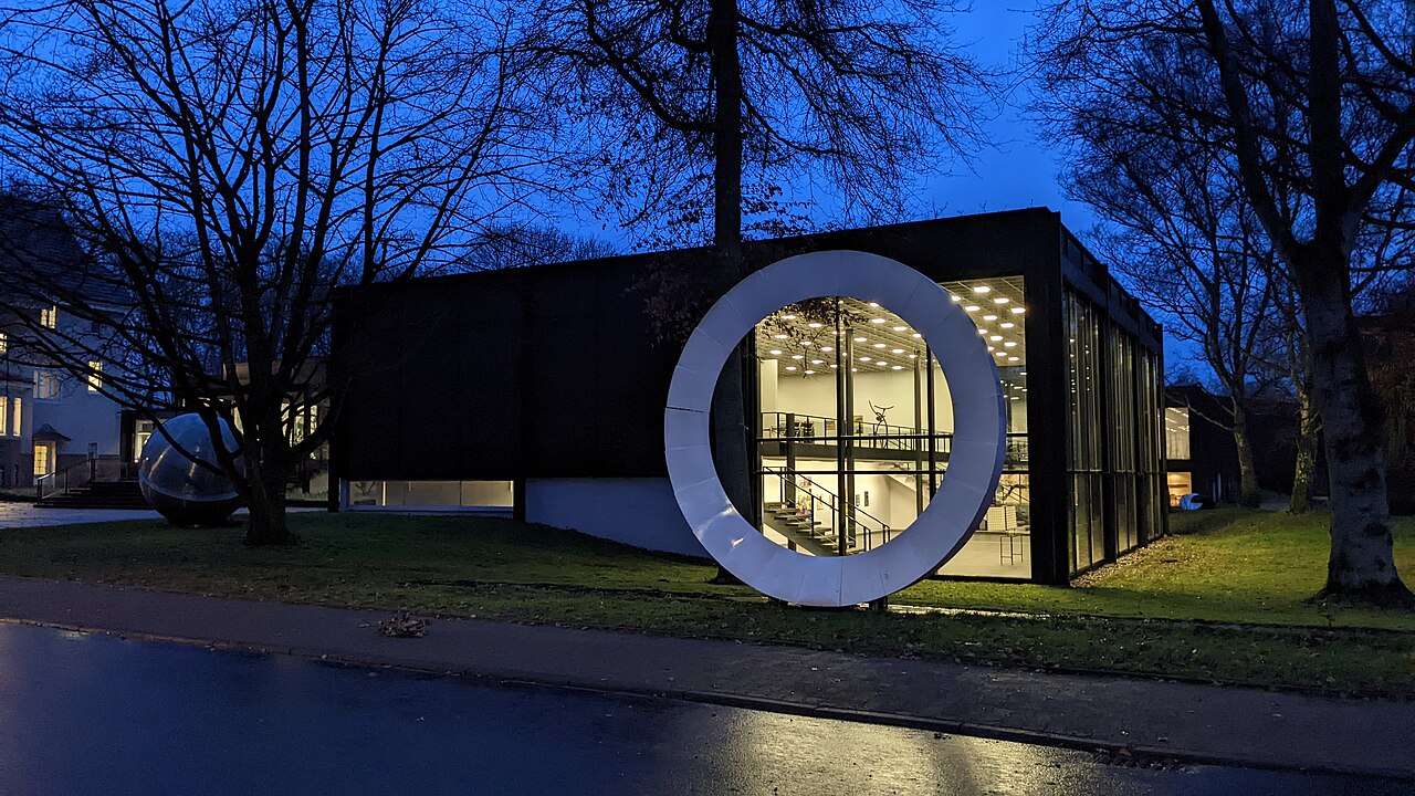 A low black glass-walled modern museum at blue dusk with a large illuminated white ring sculpture standing on the lawn.