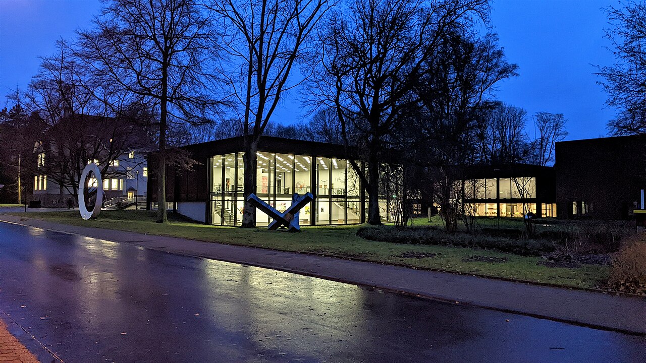 The lit museum pavilion seen from across a rain-wet street at dusk, its glass hall glowing among bare winter trees.
