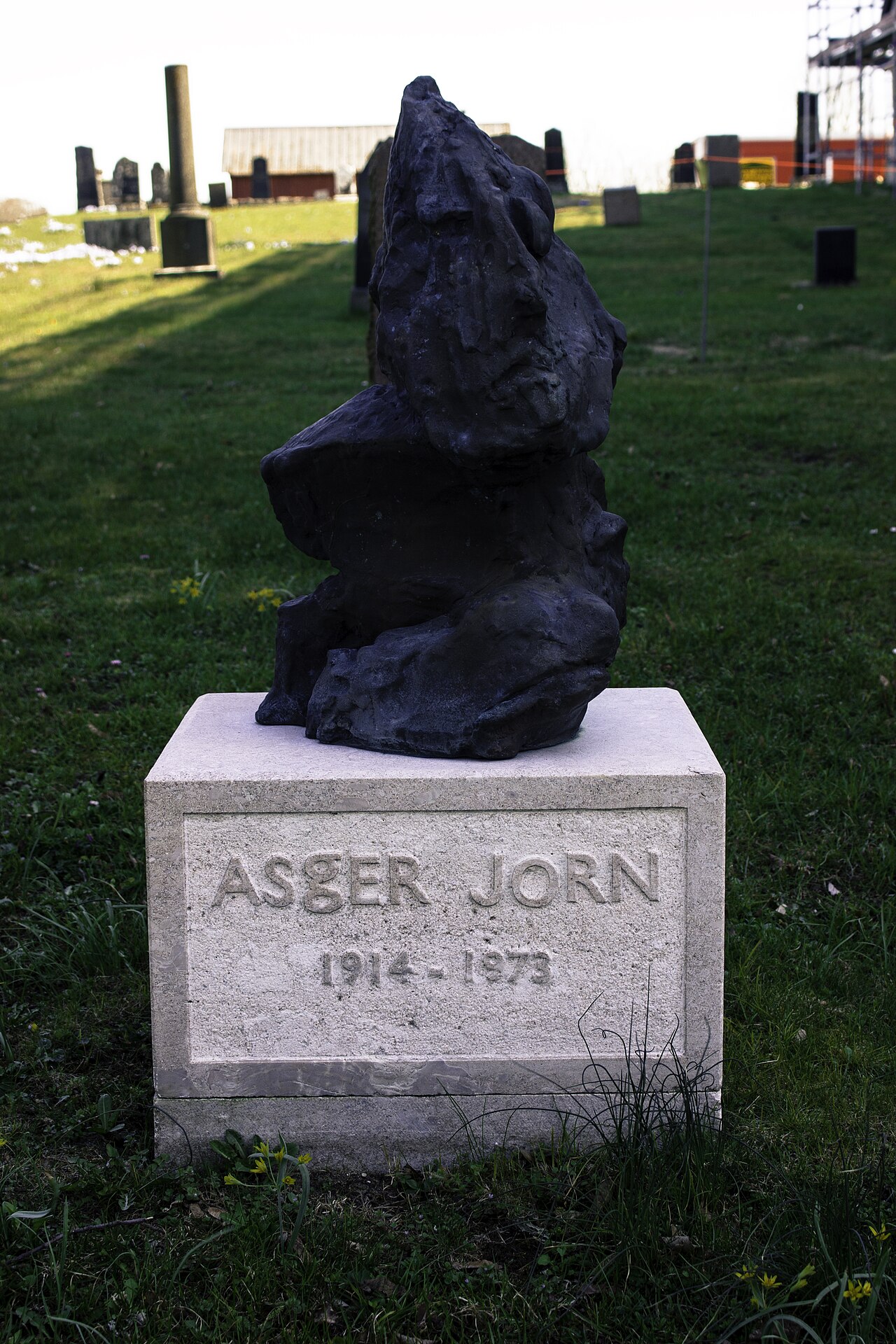 A rough, biomorphic black bronze sculpture — a crude beast-like mass — mounted on a pale stone plinth carved ASGER JORN 1914–1973, standing in a green churchyard.