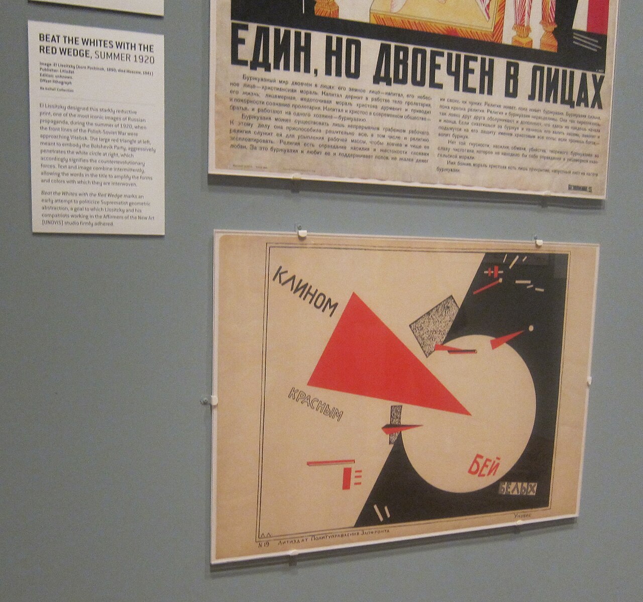 Museum display: El Lissitzky's Beat the Whites with the Red Wedge mounted beside a bold Cyrillic agitprop broadside and an English wall label explaining the work.