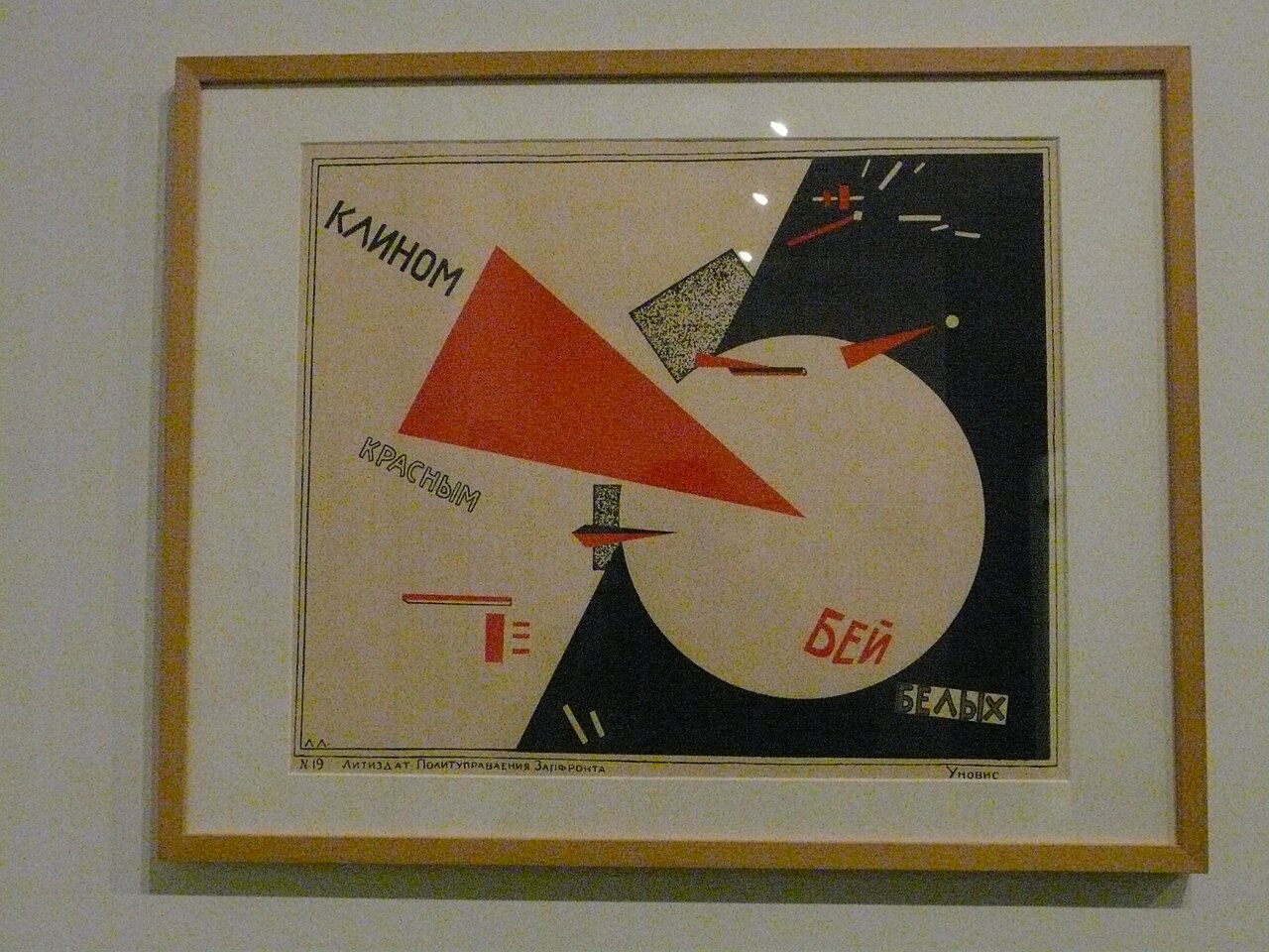A framed print of El Lissitzky's Beat the Whites with the Red Wedge hanging on a gallery wall, its red triangle rendered in a warm orange tone.