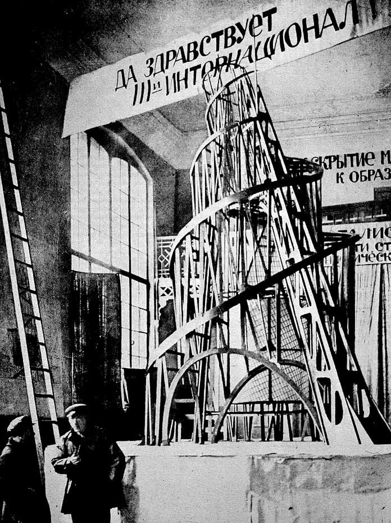 Black-and-white 1919 photograph of the wooden model for the Monument to the Third International — Tatlin's Tower — a leaning spiral lattice; Tatlin and an assistant stand beside it beneath a banner.