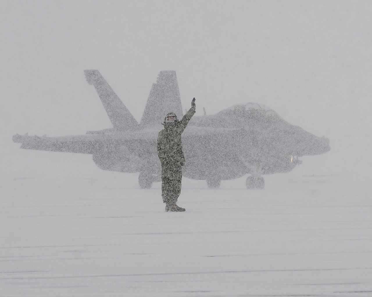 A US Navy ground crew member signalling to an EA-18G Growler aircraft during a snowstorm at Naval Air Facility Misawa, Japan.
