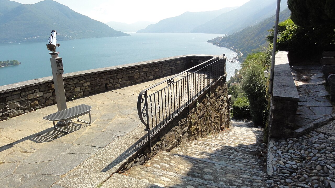A stone terrace with a railing and a viewing telescope at Ronco sopra Ascona, looking south-west down the length of Lake Maggiore between steep wooded hills.
