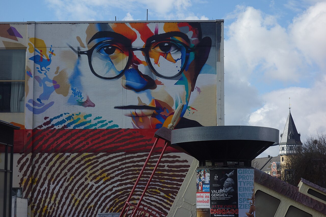 Large outdoor mural of Theodor Adorno's face rendered in vivid multicoloured brushstrokes on the side of a building in Frankfurt, with a red band and thumbprint-like patterns below, under a partly cloudy sky.