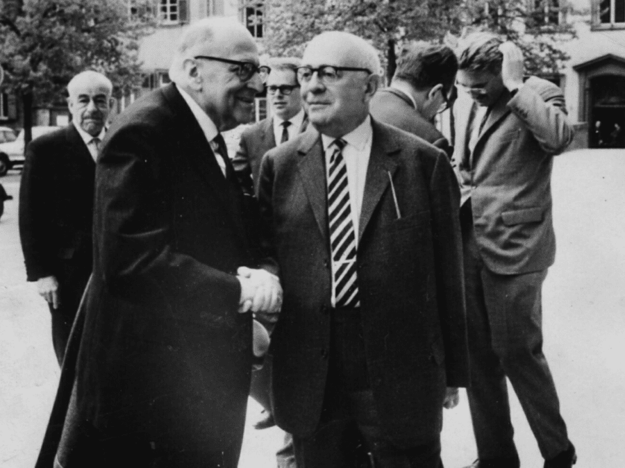 Black-and-white 1964 photograph in Heidelberg: Max Horkheimer at front left shaking hands with Theodor Adorno at front right, both in dark suits and glasses, with a young Jürgen Habermas visible in the background at right.