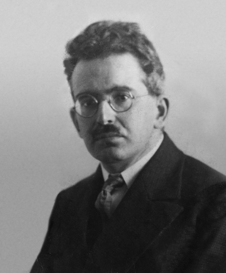 Sepia-toned 1928 portrait photograph of Walter Benjamin — bespectacled, dark-haired with a moustache, in a dark suit and patterned tie against a plain studio backdrop.