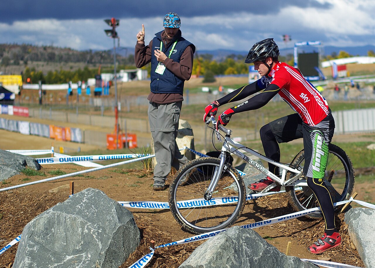A mountain-bike trials rider balances motionless on his bicycle mid-competition, body and machine held in dynamic equilibrium against gravity.
