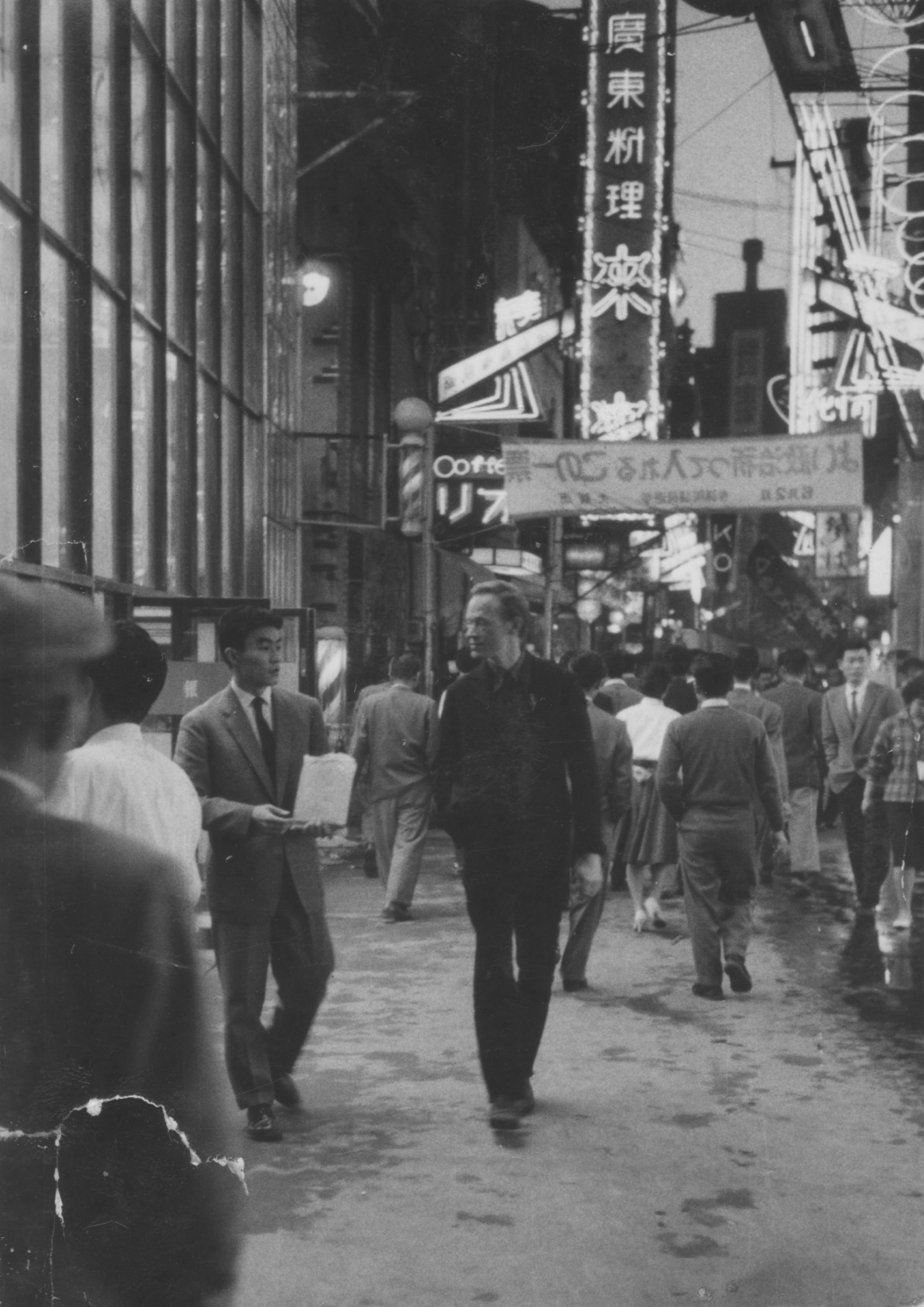 Black-and-white street photograph, Osaka 1959: pedestrians walk a wet, neon-lit shopping arcade at night beneath vertical Japanese signage; the painter Christo Coetzee and Michio Yoshihara move through the crowd.