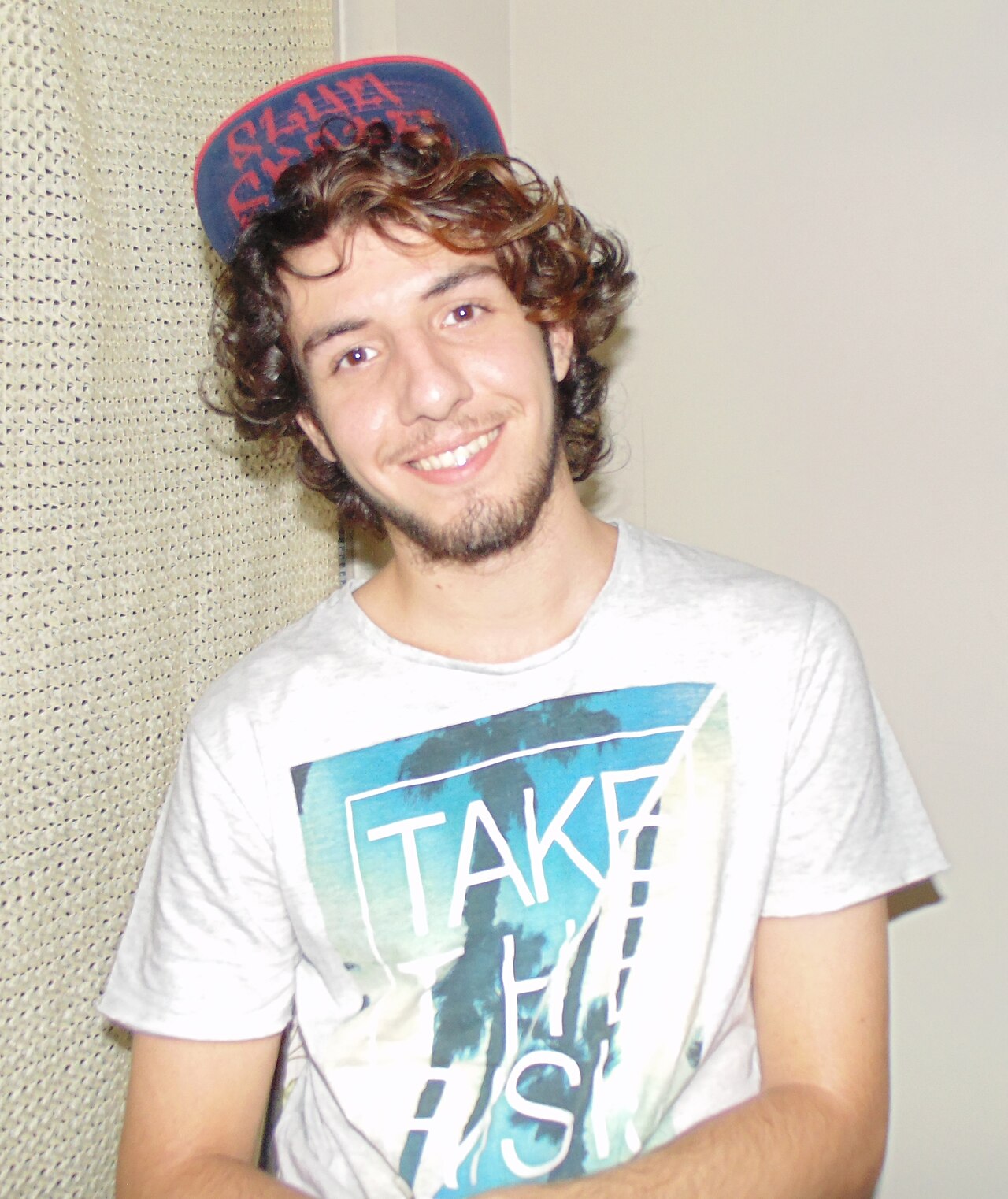 Portrait of a smiling young dance artist in a casual T-shirt and cap against a plain wall.