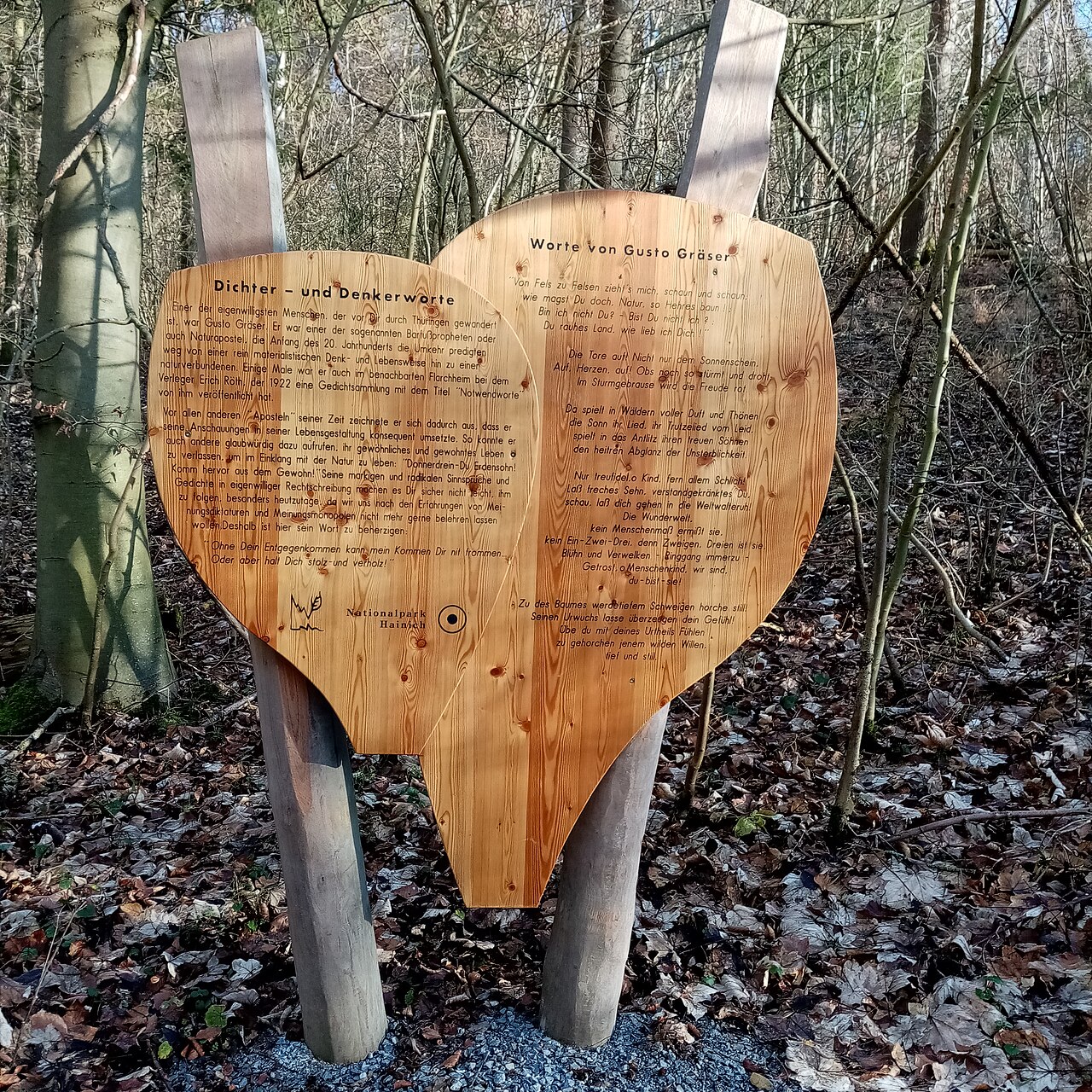 Two large heart-shaped wooden panels in a bare forest, carved with poems by Gusto Gräser, propped on log posts among fallen leaves.