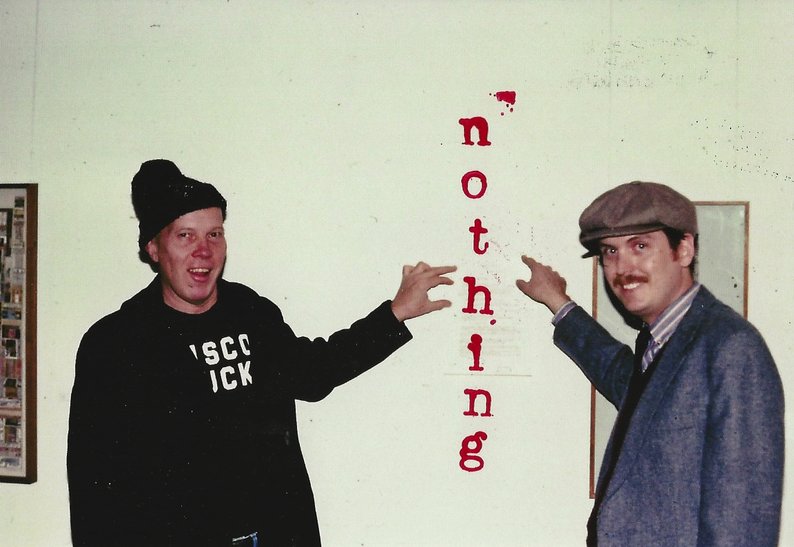 Two men in a room stand on either side of the word NOTHING rubber-stamped vertically in red on a white wall. Both grin and gesture toward the word. On the left, a man in a dark hat and sweatshirt; on the right, Ray Johnson in a flat cap and grey jacket.