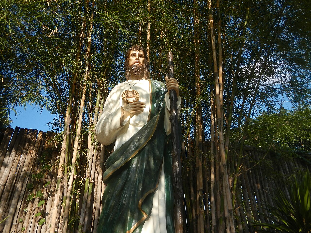 A painted outdoor statue of Saint Jude Thaddeus holding a staff amid tall bamboo, rendered in an amber-magenta duotone with scanlines.