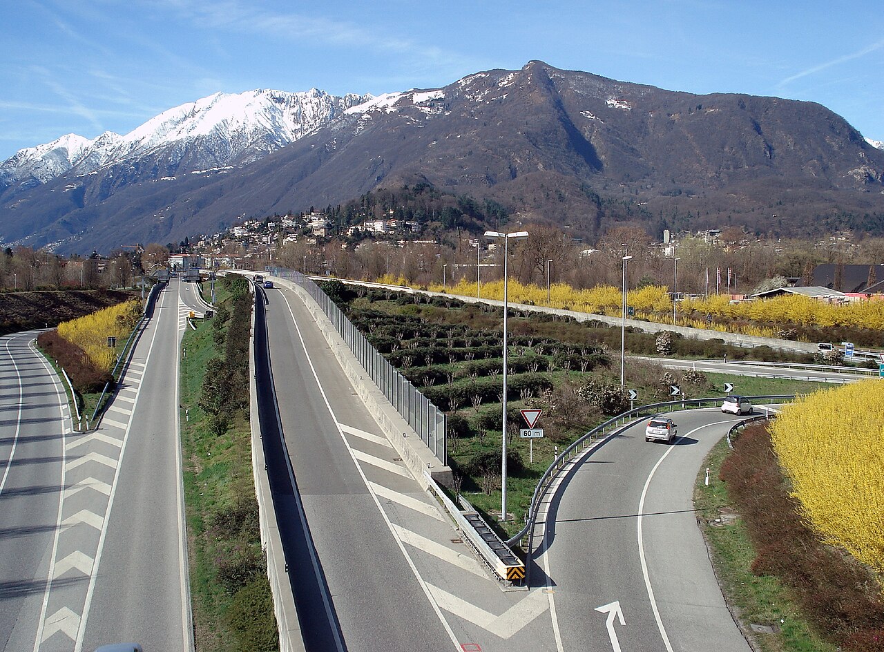 The partly-built Monte Verità hill above Ascona, seen between snow-capped alpine peaks and a modern road, flowering yellow shrubs along the verges.