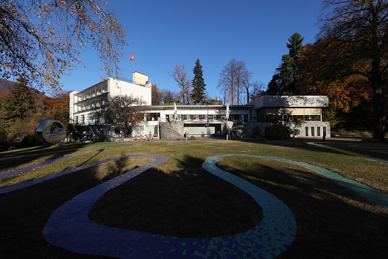 The white modernist Hotel Monte Verità in autumn, a Swiss flag on its roof, with sweeping mosaic-tiled paths curving across the lawn before it.