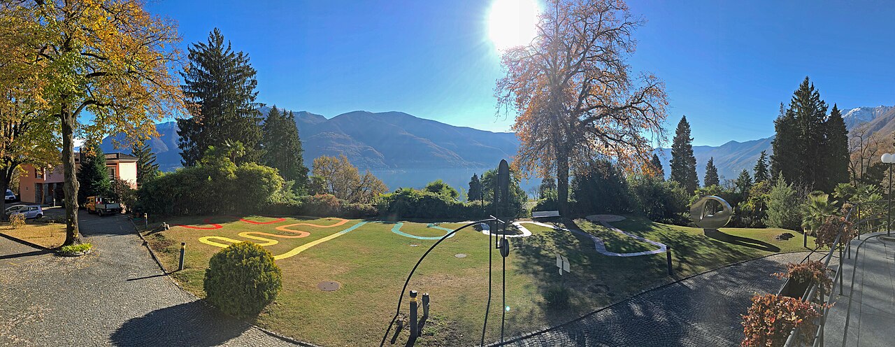 Panoramic view from the Hotel Monte Verità terrace over sunlit gardens and Lago Maggiore, mountains ringing the far shore under a bright sun.