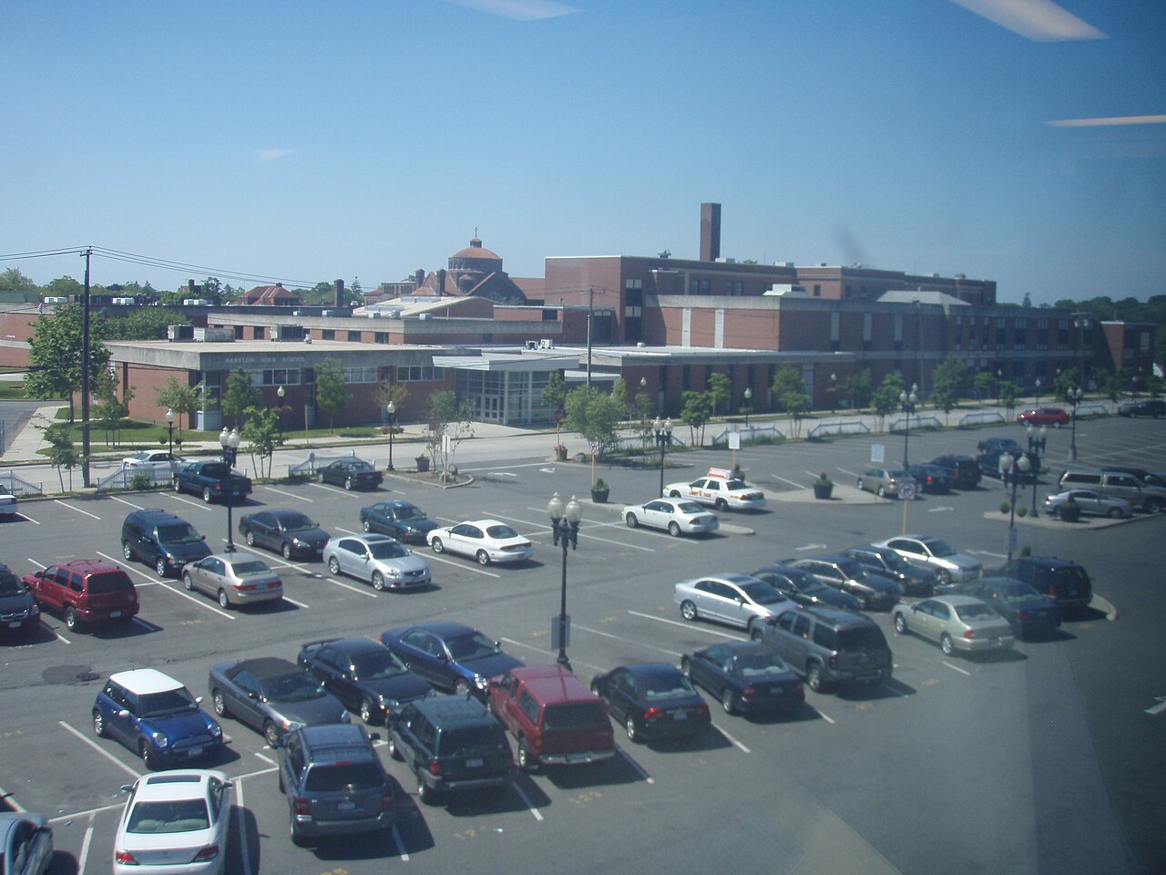 Aerial view of Babylon High School, Babylon, New York: a low brick school building fronted by a large asphalt parking lot filled with parked cars.