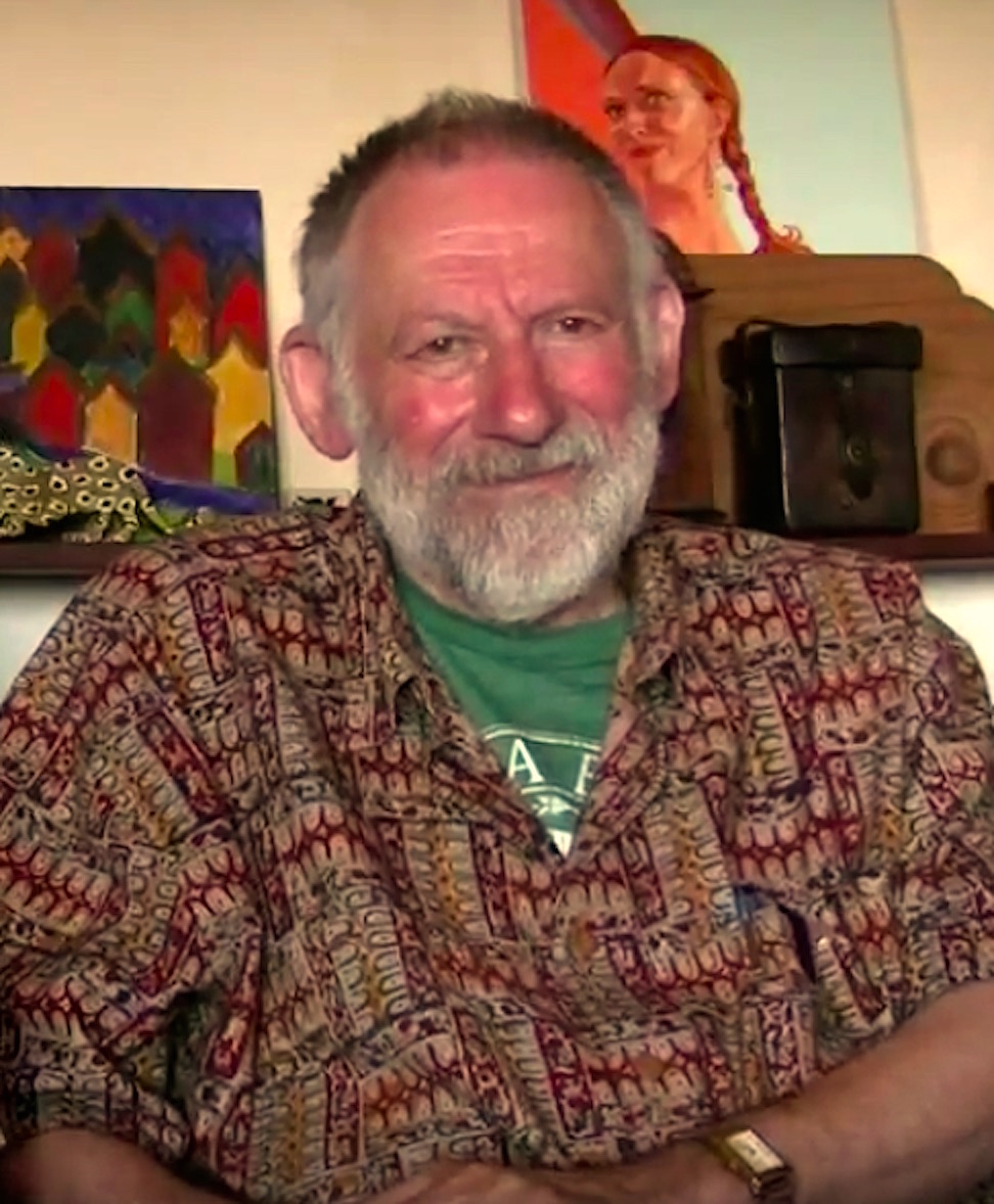 Christopher Alexander in a patterned shirt, seated before paintings and a wooden radio, smiling gently.