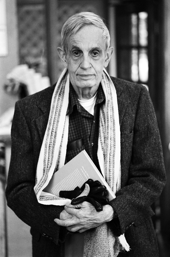 Black-and-white portrait of mathematician and economist John Forbes Nash Jr. in later life, wearing a tweed jacket and striped scarf, holding a book, photographed indoors.