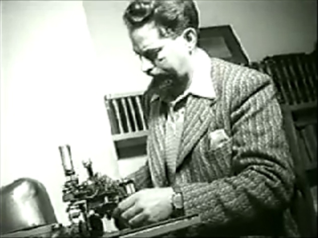Grainy black-and-white still of W. Grey Walter, bearded and in a tweed jacket, seated at a bench adjusting a small electromechanical apparatus.