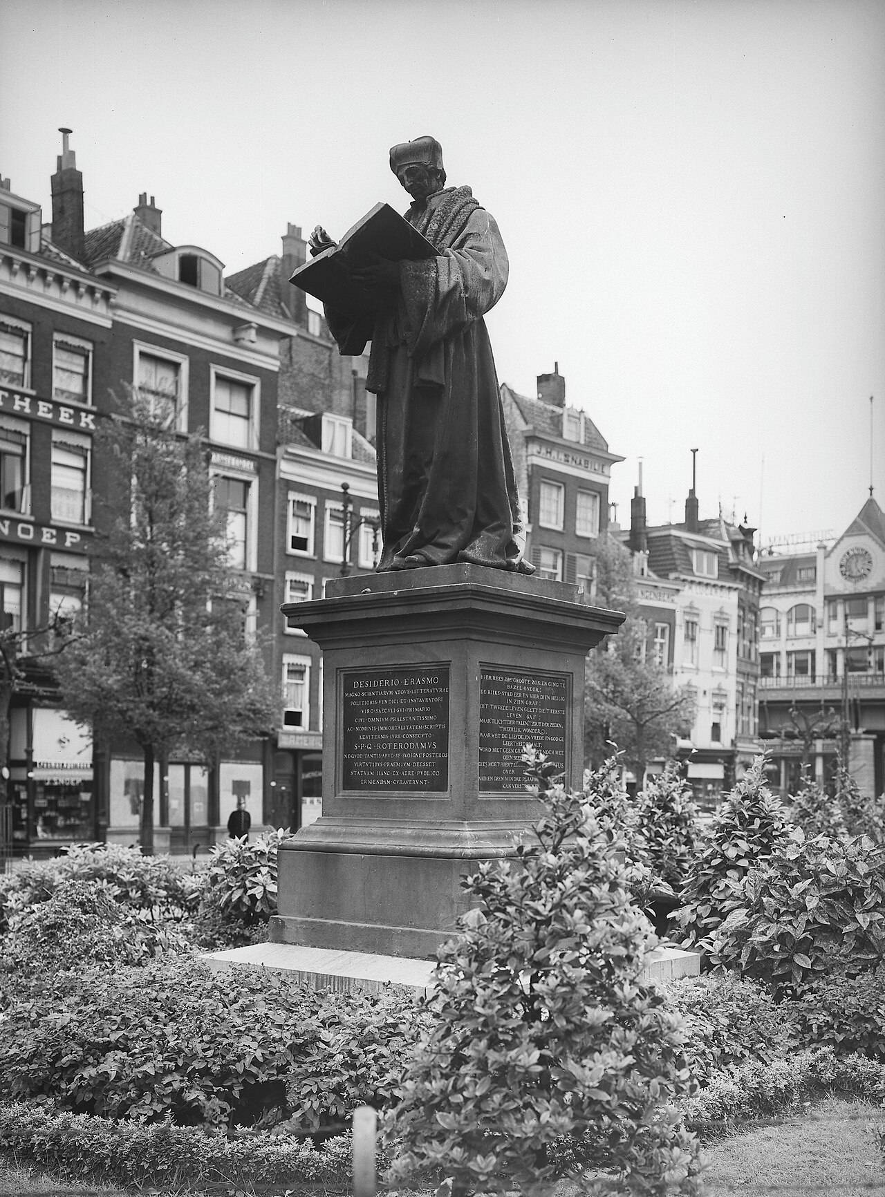 Bronze statue of Erasmus reading an open book, standing on a Latin-inscribed plinth in a public square in Rotterdam.