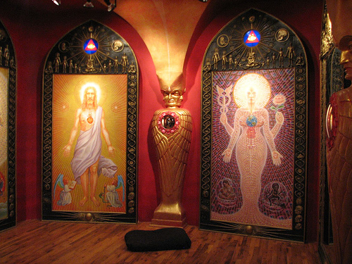 Interior of a chapel with deep-red walls, a gilded winged sculpture, and two arched panels depicting luminous haloed figures.
