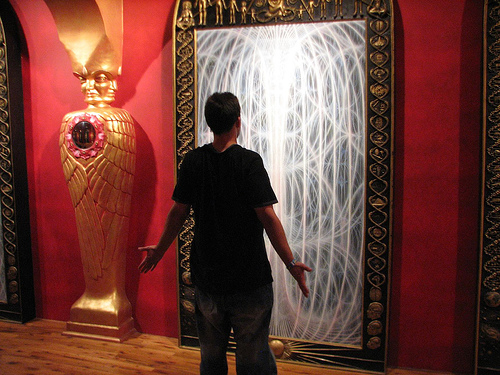 A visitor stands with arms open before a large luminous panel of white filament-like light, beside a golden winged figure on a red wall.