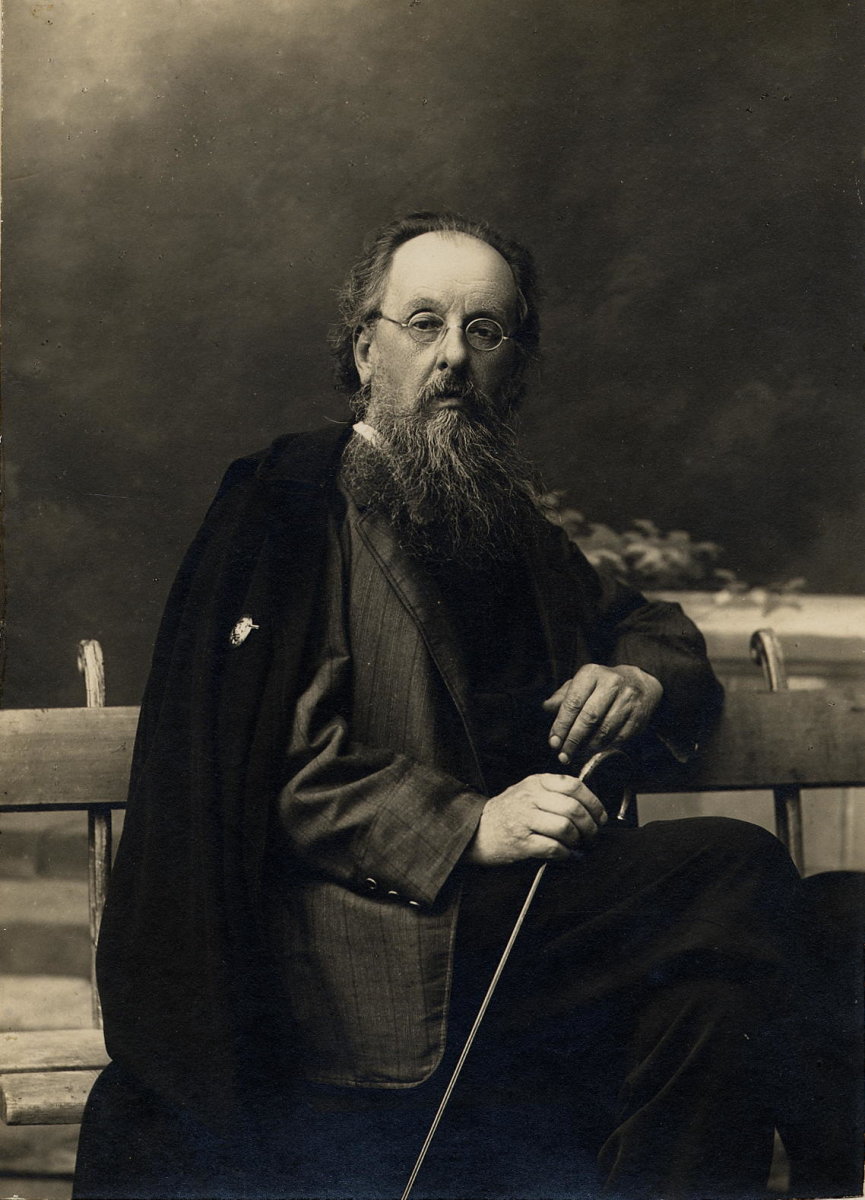 Sepia studio portrait of Konstantin Tsiolkovsky, an elderly bearded man in round spectacles seated on a bench with a cane, photographed against a painted backdrop.