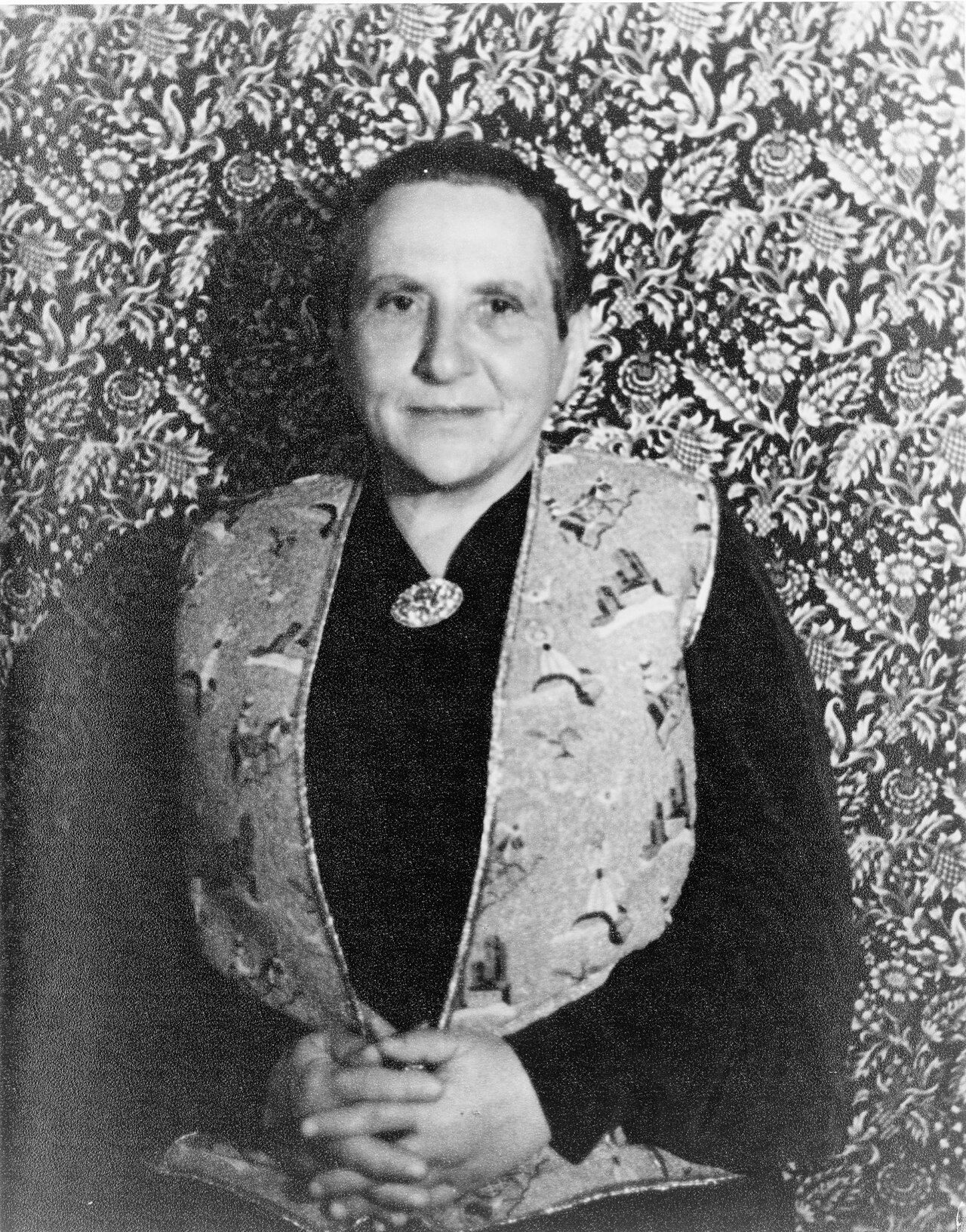 Photographic portrait of Gertrude Stein seated before a densely patterned floral-damask wallpaper, wearing a dark robe with an embroidered collar, hands folded.