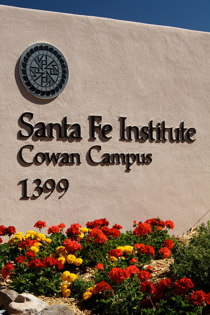 The Santa Fe Institute main gate: a rounded adobe wall bearing a stone medallion and the raised letters 'Santa Fe Institute · Cowan Campus · 1399', with red and yellow desert flowers below against a deep blue sky.