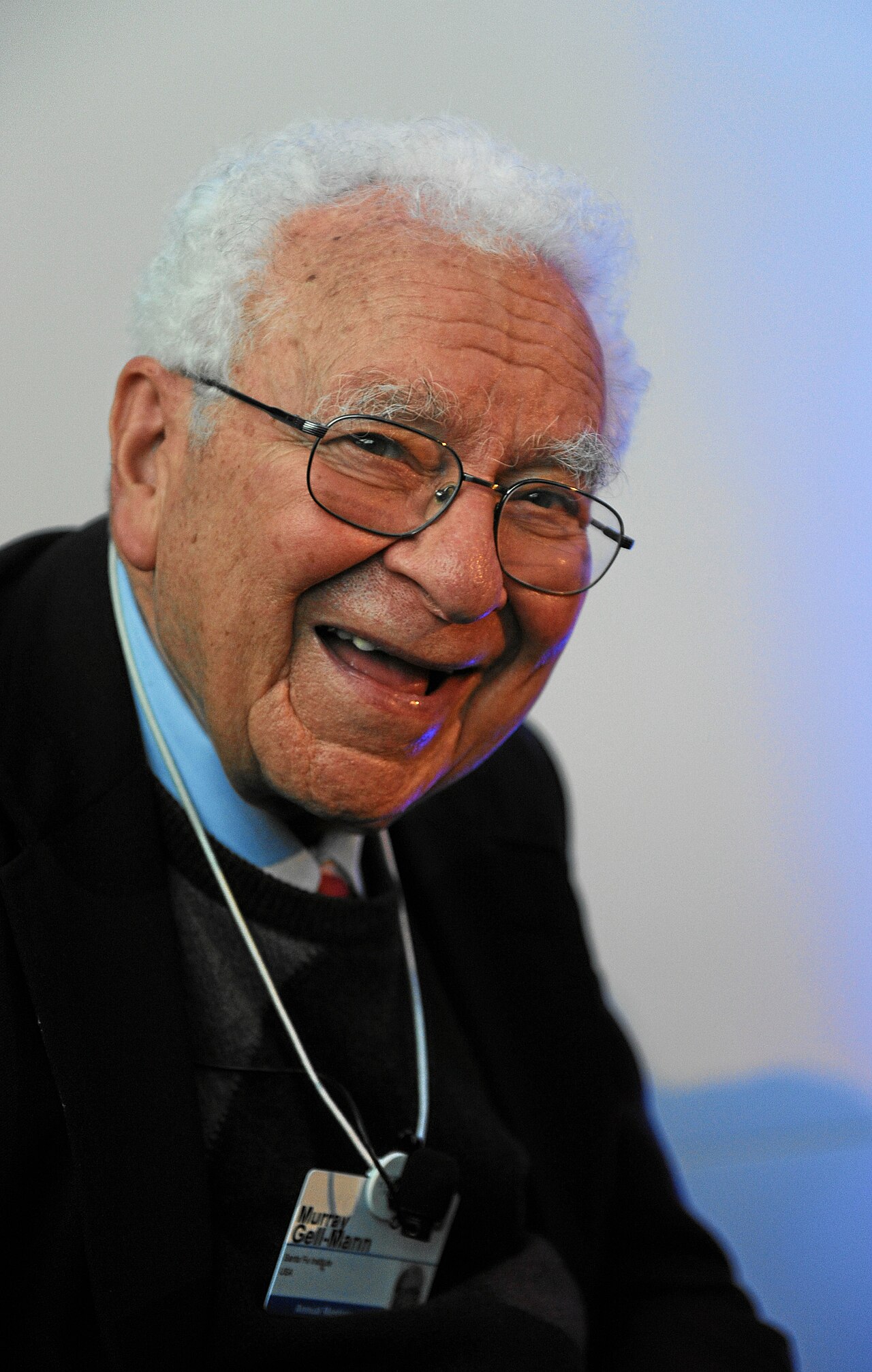 Physicist Murray Gell-Mann, silver-haired and grinning, wearing round glasses and a Santa Fe Institute conference badge, photographed at the 2012 World Economic Forum.