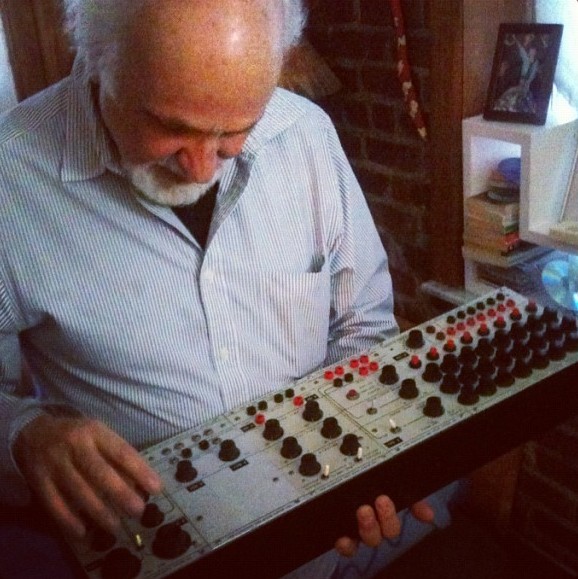 Morton Subotnick, white-haired and bearded, looks down intently at a vintage 1960s Buchla 100 module panel held across his lap, its rows of black and red knobs catching the light in a warm brick-walled room.