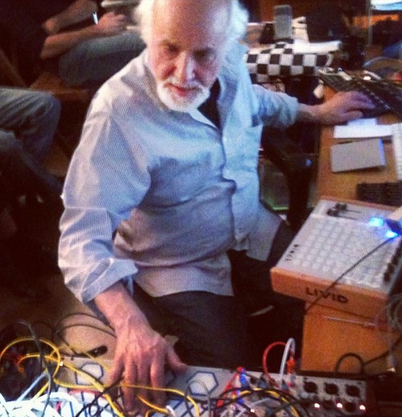 Morton Subotnick sits on the floor before a modular synthesizer, hands over a tangle of yellow and red patch cables, an audience seated close around him in a dim room.