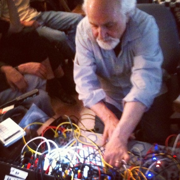 Subotnick leans over a modular synthesizer thick with colored patch cords and glowing blue indicator lights, reaching in to change a connection mid-performance.