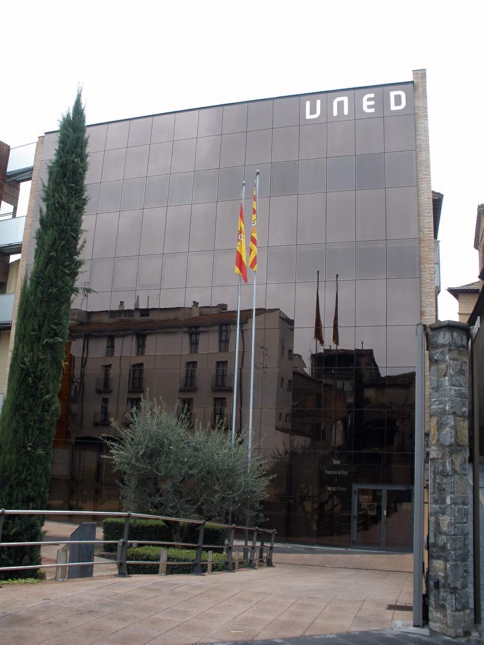 A modern dark-glass building lettered UNED, with two flagpoles and a slender cypress tree, reflecting older stone houses in its facade.