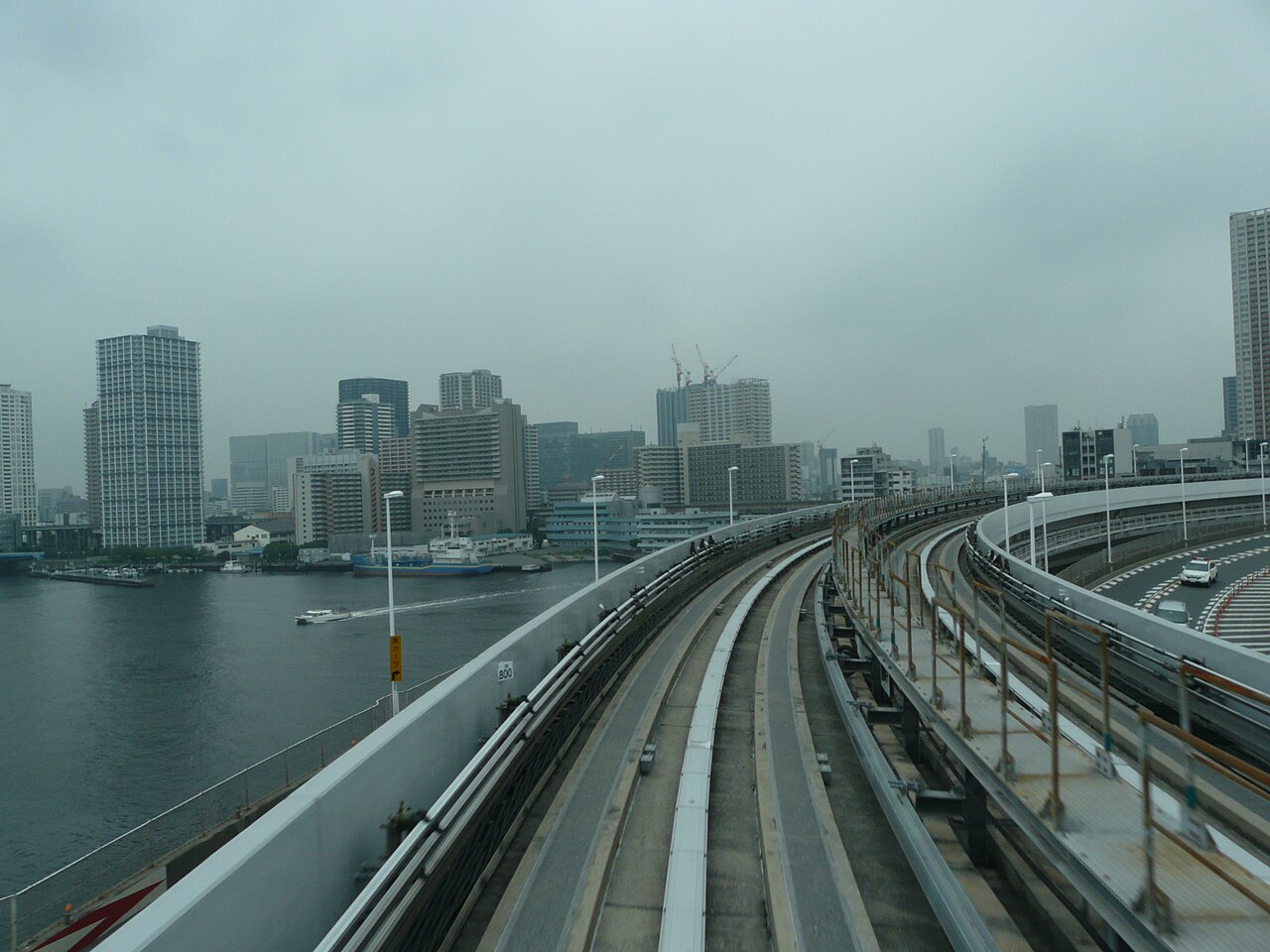 A curving elevated transit track sweeps away into a flat grey industrial cityscape beside grey water — the endless road, receding.