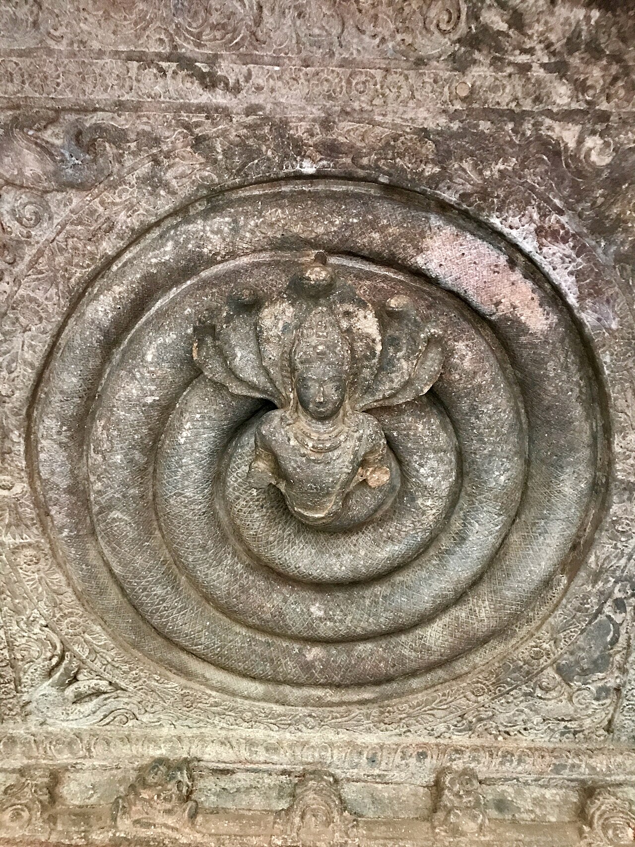 A stone relief of a deity seated within the tightly coiled loops of a serpent, carved into an ancient cave ceiling — a spiral turning inward toward its centre.
