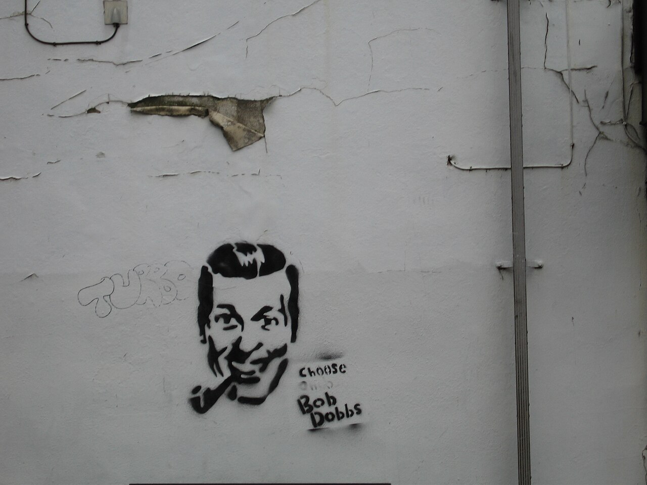 A black stencil graffito of J.R. "Bob" Dobbs — a grinning 1950s clip-art everyman with a pipe — sprayed on a cracked white wall, captioned 'Choose Bob Dobbs.'