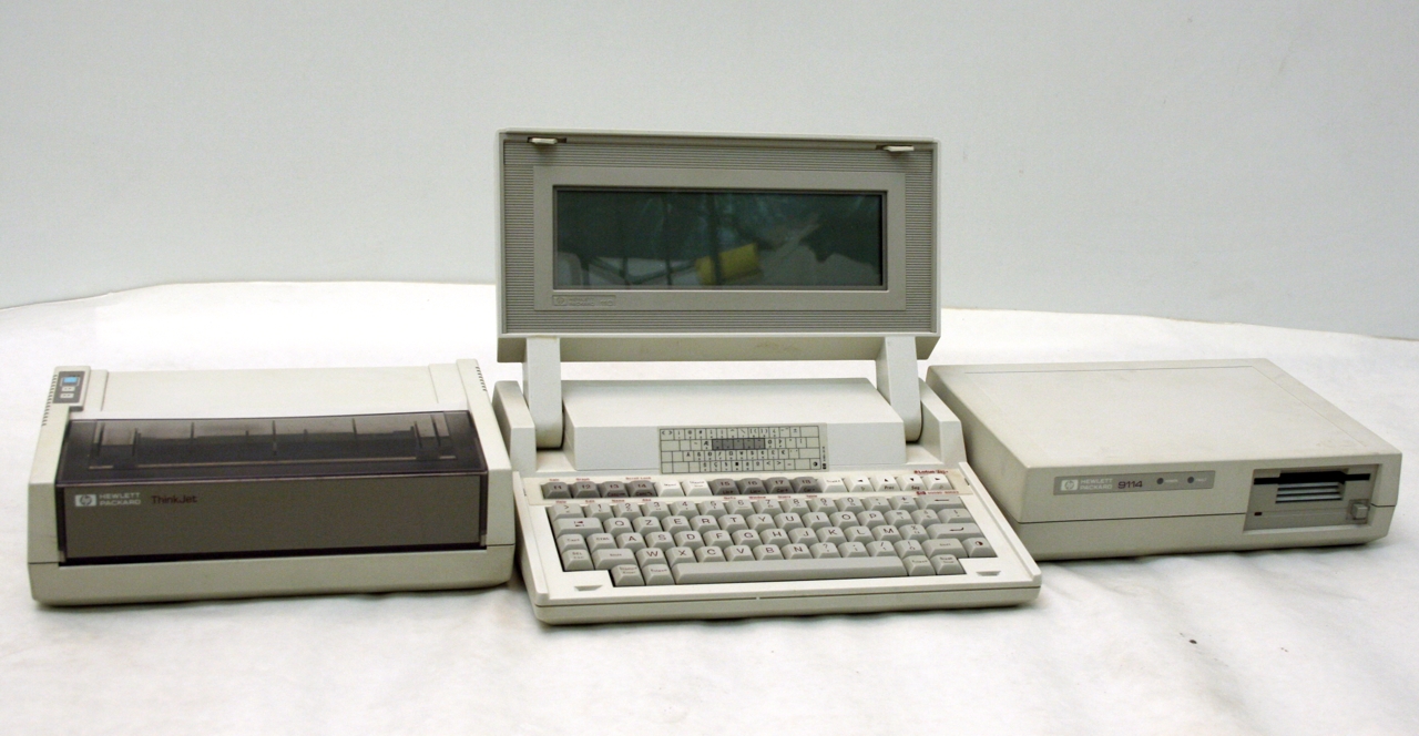 An open beige 1980s Hewlett-Packard portable computer with a narrow flip-up LCD screen and a full keyboard, flanked by a ThinkJet printer and a disk-drive unit.