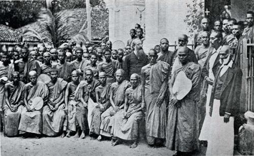 A sepia photograph, 1883: the bearded, white-haired Colonel Henry Steel Olcott in a Western suit standing among rows of seated and standing Buddhist monks outside a temple in Colombo, Ceylon.