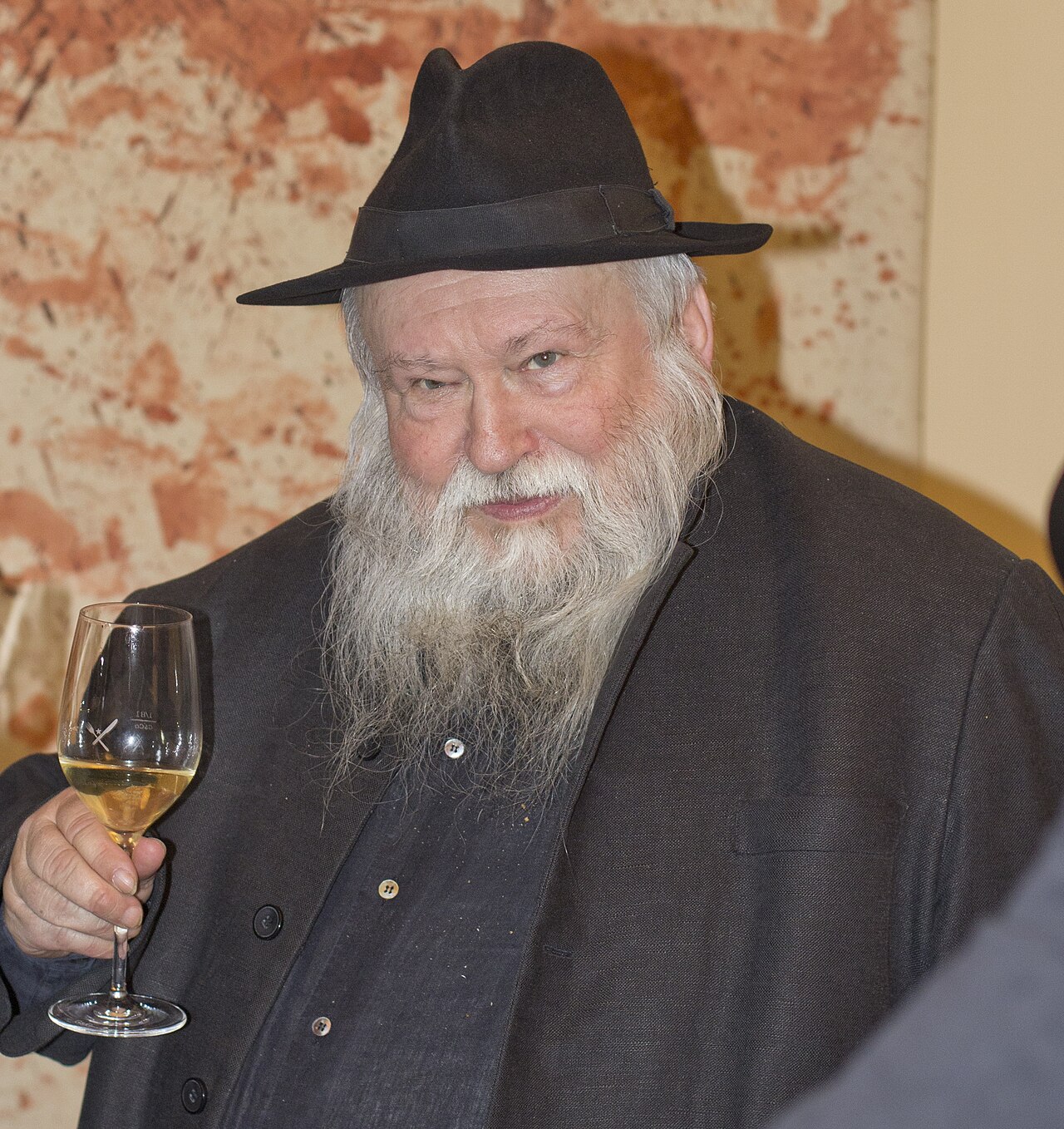 Hermann Nitsch as an old man, in a black hat and dark coat, holding a glass of wine before one of his blood-red splatter paintings.
