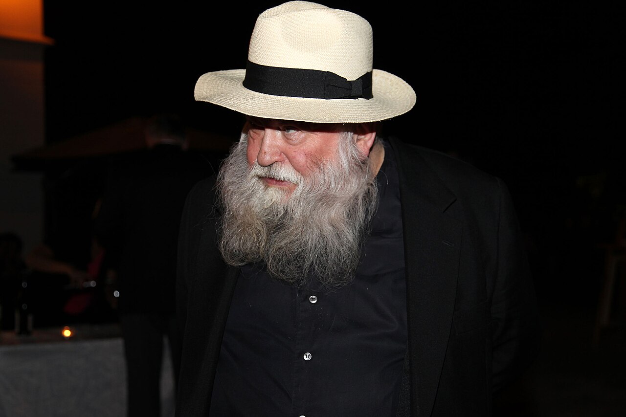 Hermann Nitsch at night in a pale hat and black clothing, emerging from darkness at a festival in 2010.